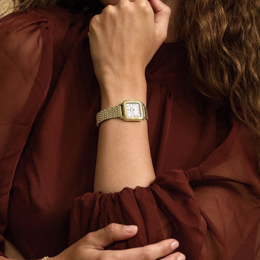 A woman's torso and arm. She is wearing a chic burgundy blouse and a gold toned square Cluse watch
