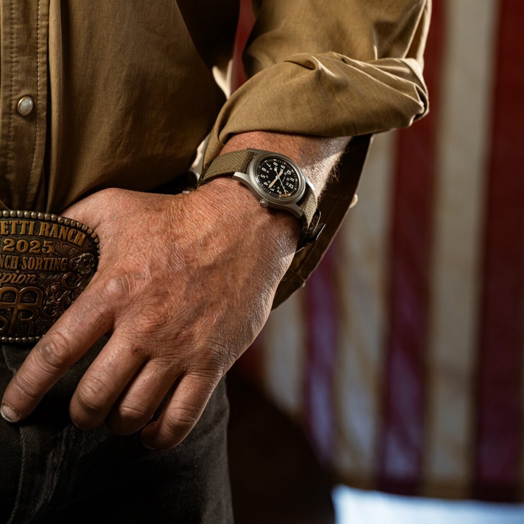 A man's hand, thumb behind belt buckle, wearing khaki clothes and wearing a Hamilton Khaki watch