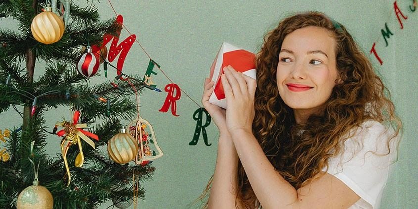 Mädchen schüttelt ein verpacktes Geschenk neben einem Weihnachtsbaum