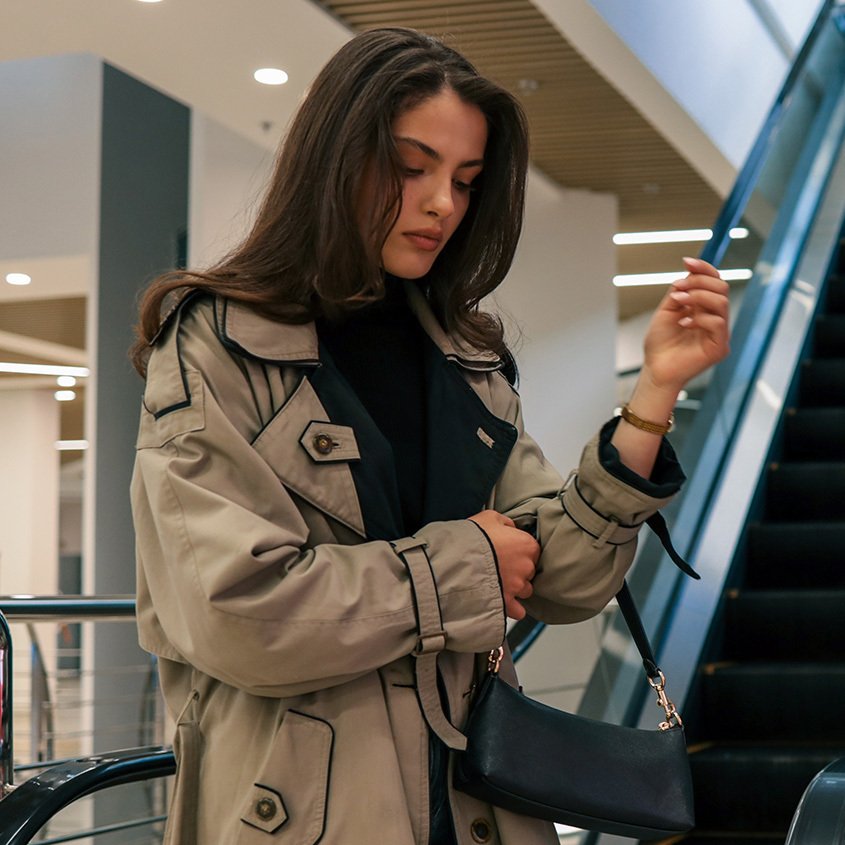 Young woman with long brown hair dressed in a trenchcoat carrying a small leather handbag