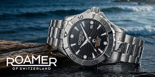 Steel Roamer dive watch with a black dial, lying on a rock with the sea behind it